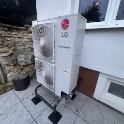 AQUAPULS SPÓŁKA Z OGRANICZONĄ ODPOWIEDZIALNOŚCIĄ - Biała pompa ciepła LG Therma V, zamontowana na zewnątrz budynku, widoczna na tle kamiennej ściany i okna. Ujęcie z niskiego kąta, podkreślające nowoczesny design urządzenia.