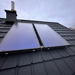 AQUAPULS SPÓŁKA Z OGRANICZONĄ ODPOWIEDZIALNOŚCIĄ - Dwa panele słoneczne zamontowane na ciemnym, spadzistym dachu z dachówki, widoczne z dołu pod kątem. W tle komin i fragmenty drzew.