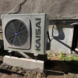 AQUAPULS SPÓŁKA Z OGRANICZONĄ ODPOWIEDZIALNOŚCIĄ - Szara jednostka zewnętrzna klimatyzacji Kaisai zamontowana na betonowych podstawach przy ścianie budynku, z widocznym przewodem elektrycznym.