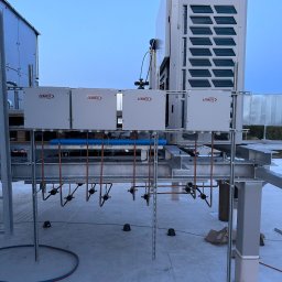 AQUAPULS SPÓŁKA Z OGRANICZONĄ ODPOWIEDZIALNOŚCIĄ - Instalacja klimatyzacji Lennox na dachu budynku, widoczne cztery skrzynki sterownicze i jednostka zewnętrzna. Miedziane rury i metalowa konstrukcja wsporcza.