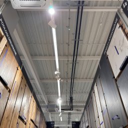 AQUAPULS SPÓŁKA Z OGRANICZONĄ ODPOWIEDZIALNOŚCIĄ - Kasetonowa klimatyzacja sufitowa Lennox w markecie budowlanym, widok z dołu na instalacje i oświetlenie, panele podłogowe po bokach.