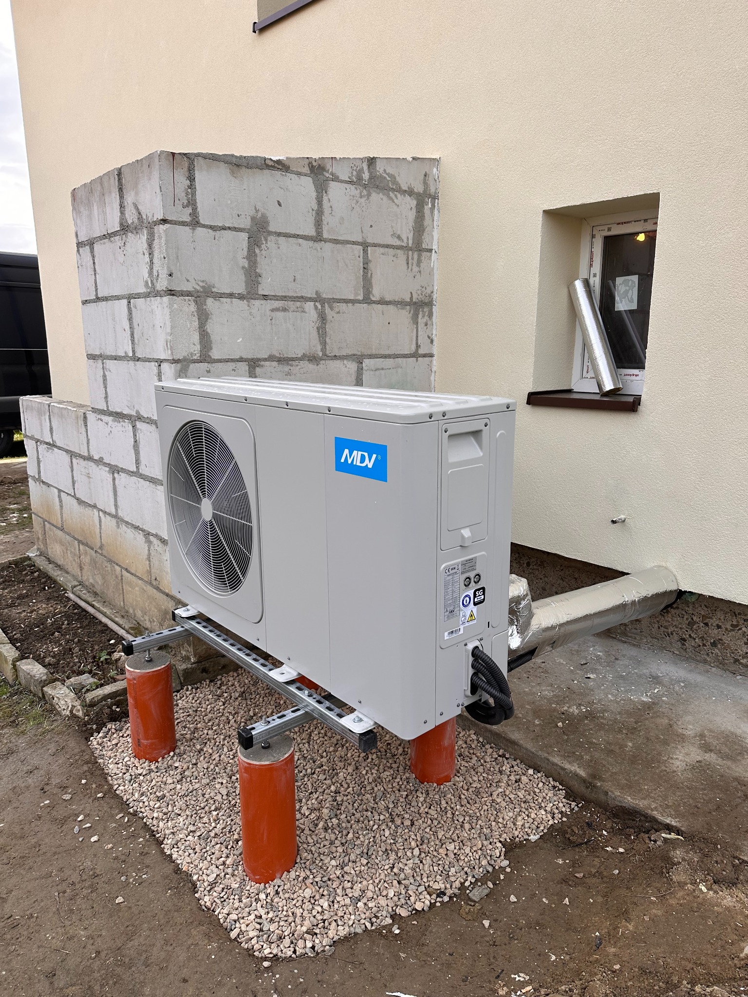 Szara jednostka zewnętrzna klimatyzacji MDV na pomarańczowych wspornikach, z widocznymi przewodami i fragmentem elewacji budynku w tle.