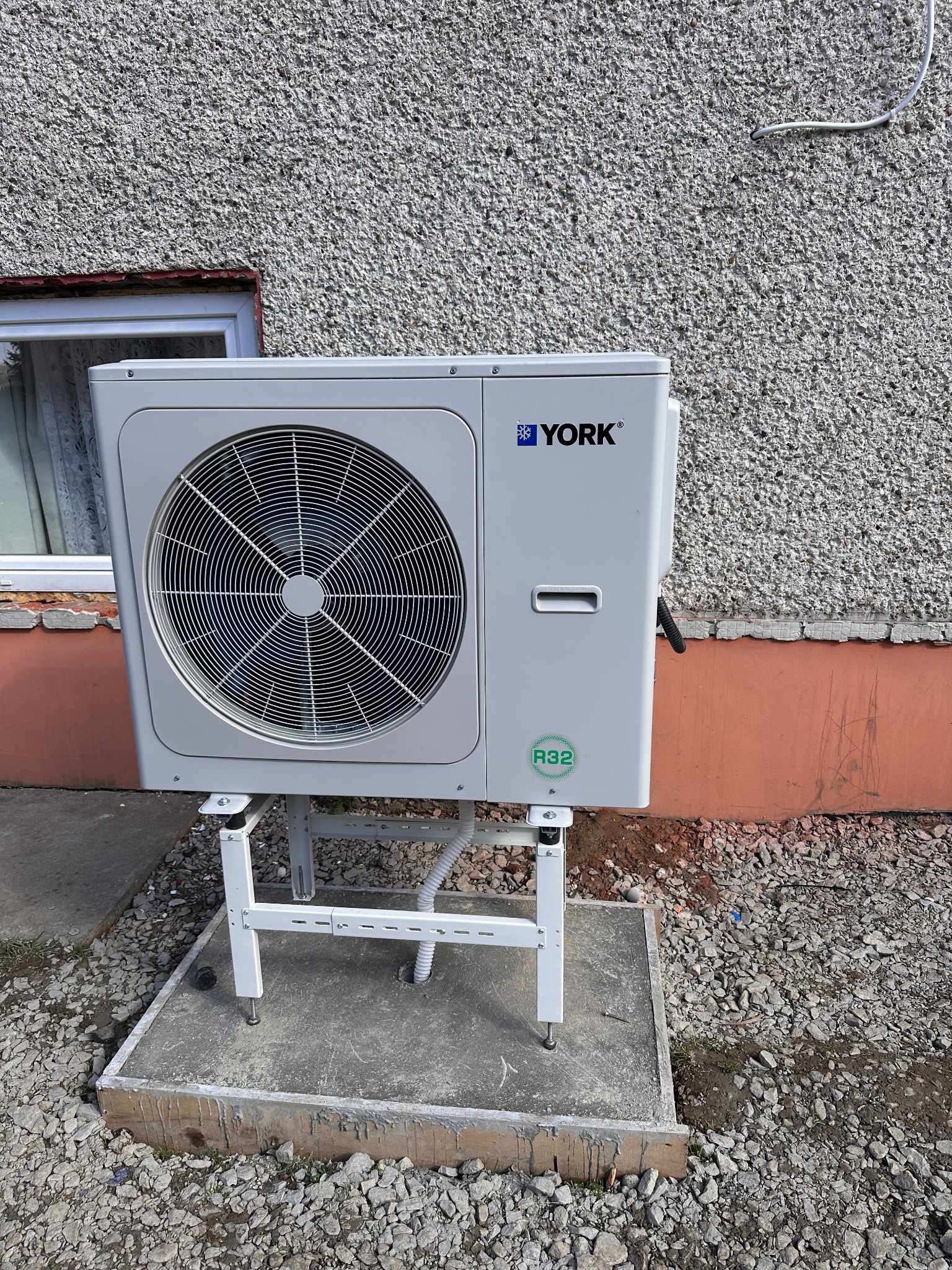 Biała jednostka zewnętrzna klimatyzacji York na betonowym fundamencie, zamontowana przy ścianie budynku o szarej, chropowatej fakturze. Widoczny zielony symbol R32.