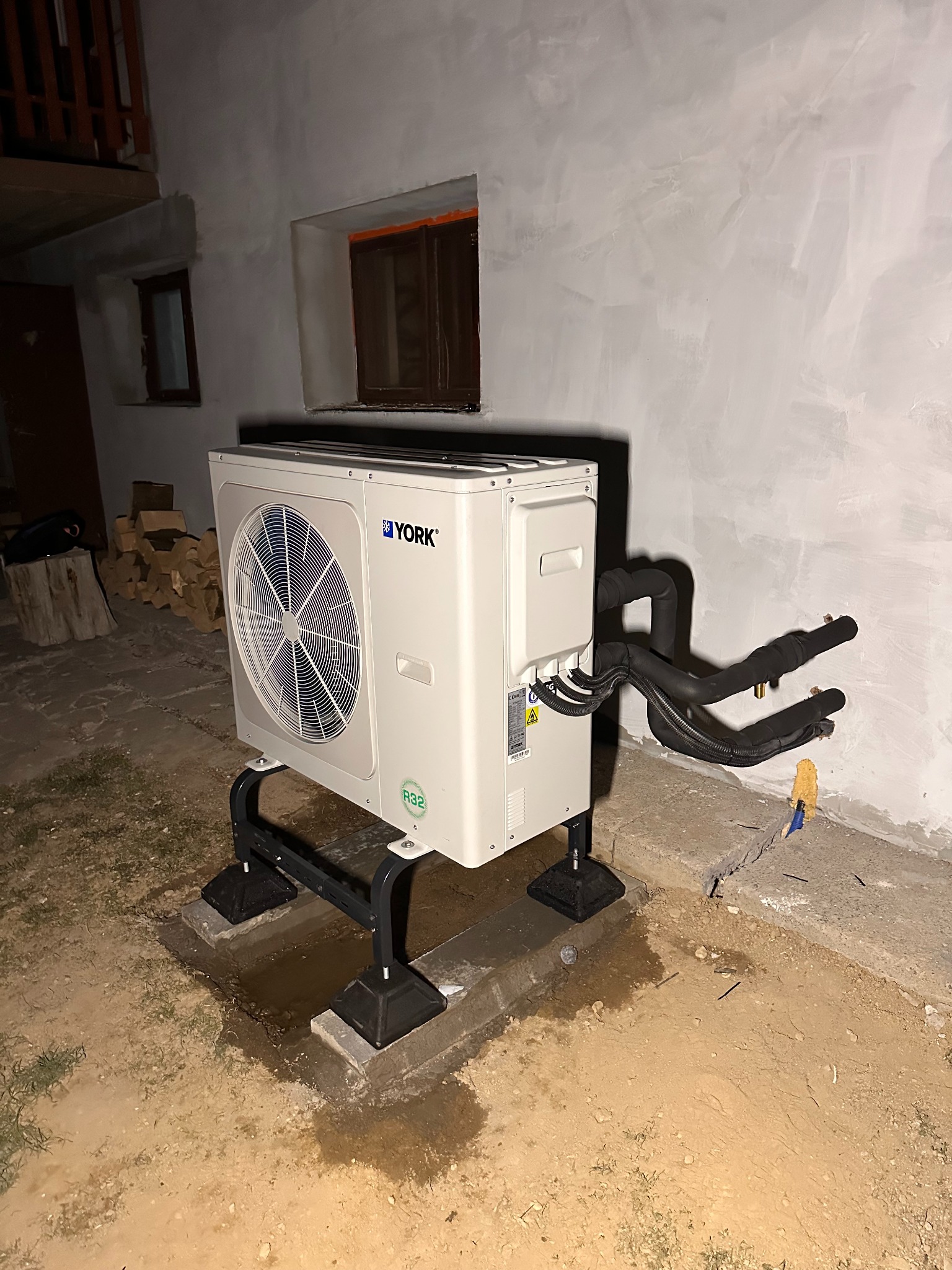 Biała pompa ciepła York na czarnej podstawie, z widocznymi rurami, zainstalowana przy szarej ścianie budynku z małymi oknami. Widoczne ślady po montażu.