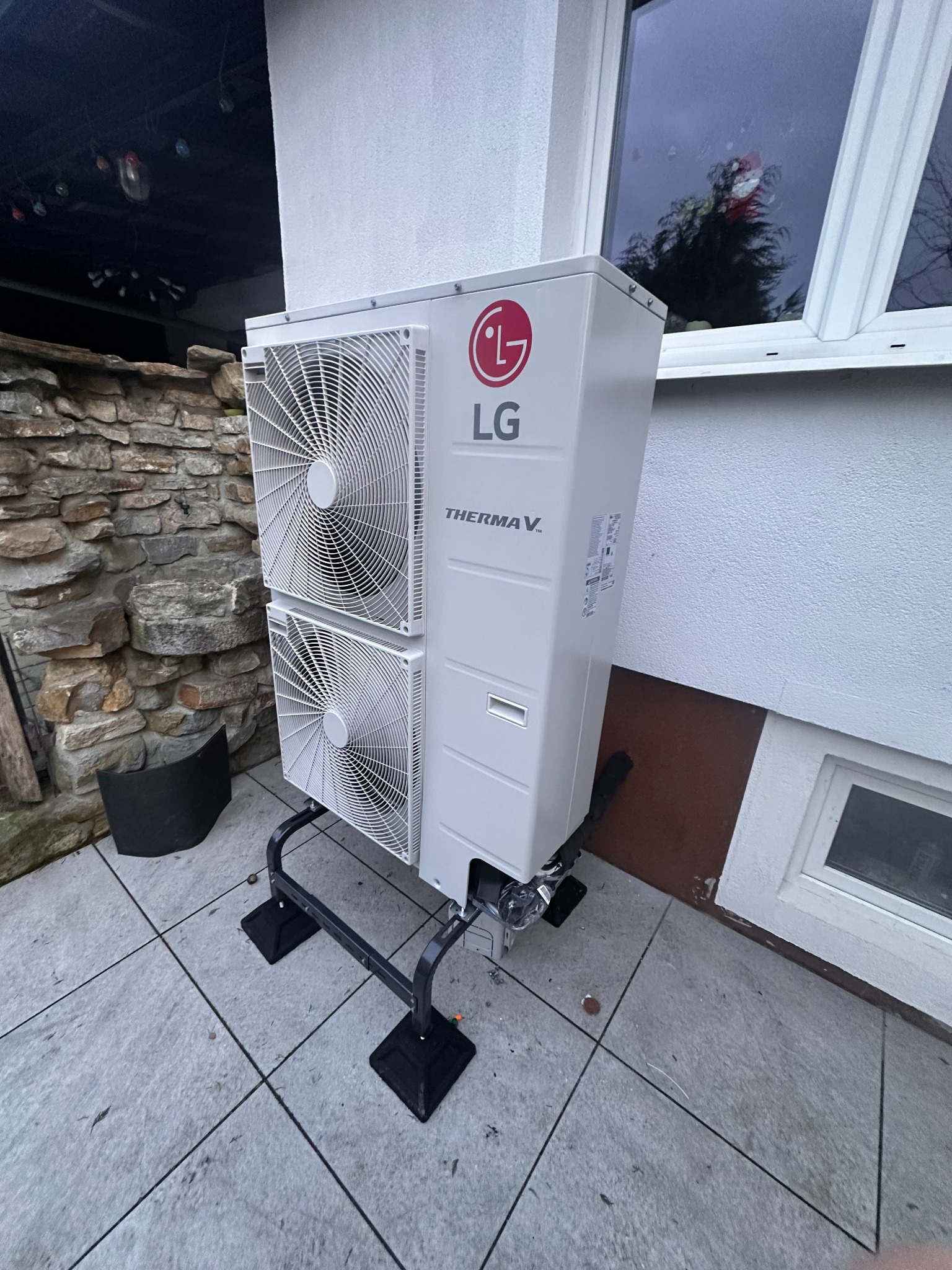 Biała pompa ciepła LG Therma V, zamontowana na zewnątrz budynku, widoczna na tle kamiennej ściany i okna. Ujęcie z niskiego kąta, podkreślające nowoczesny design urządzenia.