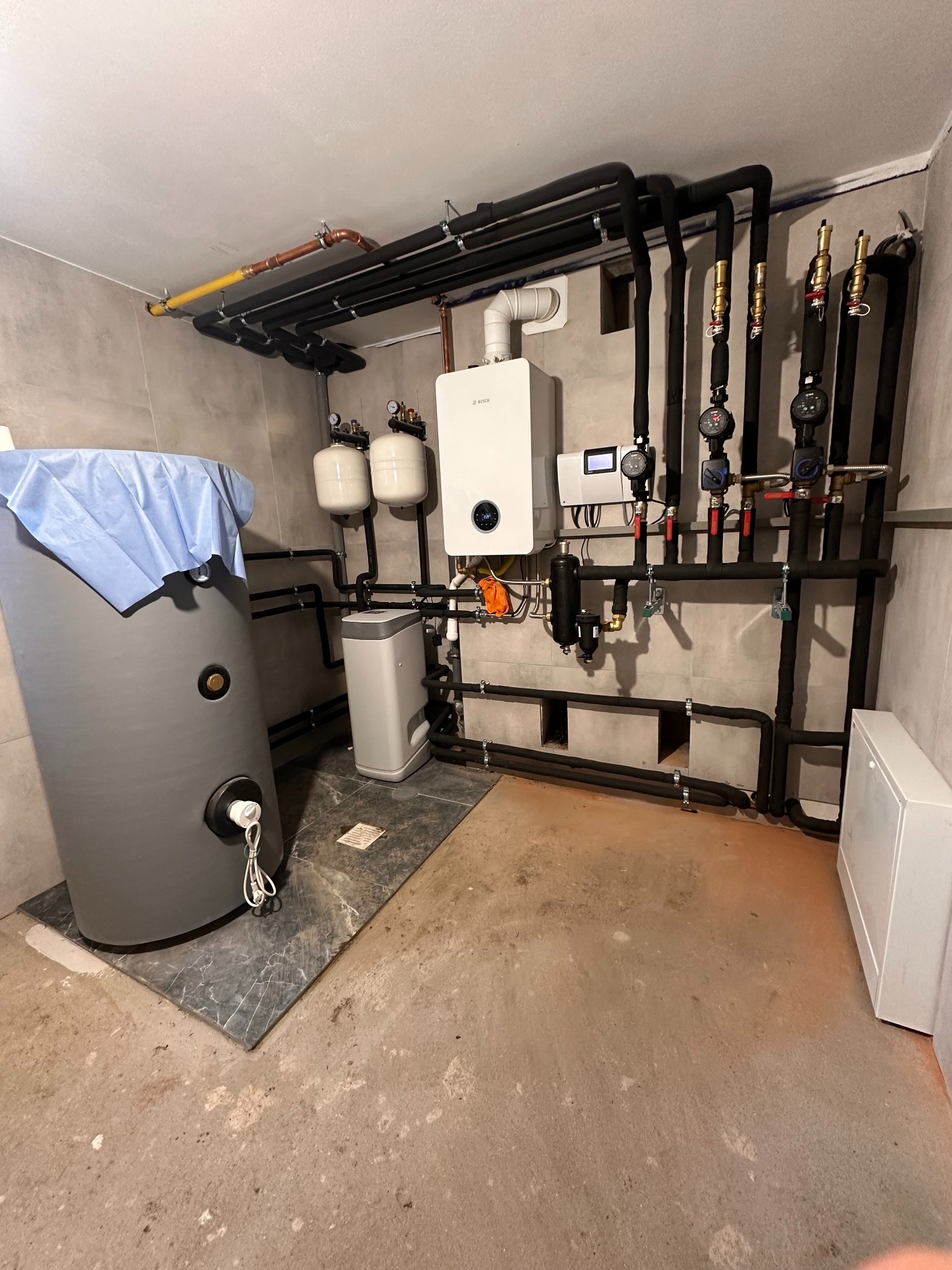 Zmontowana instalacja grzewcza z piecem gazowym Bosch, zbiornikiem i rurami w piwnicy. Widoczne elementy instalacji i podłączenia.