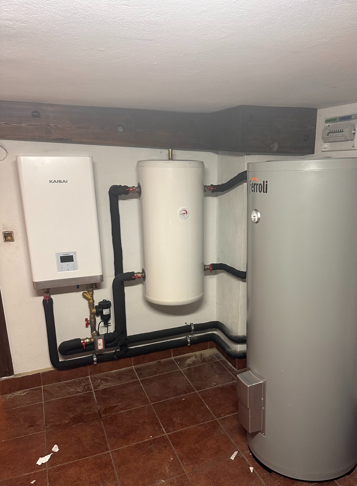 Instalacja grzewcza: biały piec Kaisai, zbiornik i szary zasobnik Ferroli, połączone rurami w izolacji. Widoczne elementy instalacji i brązowa podłoga z płytek.