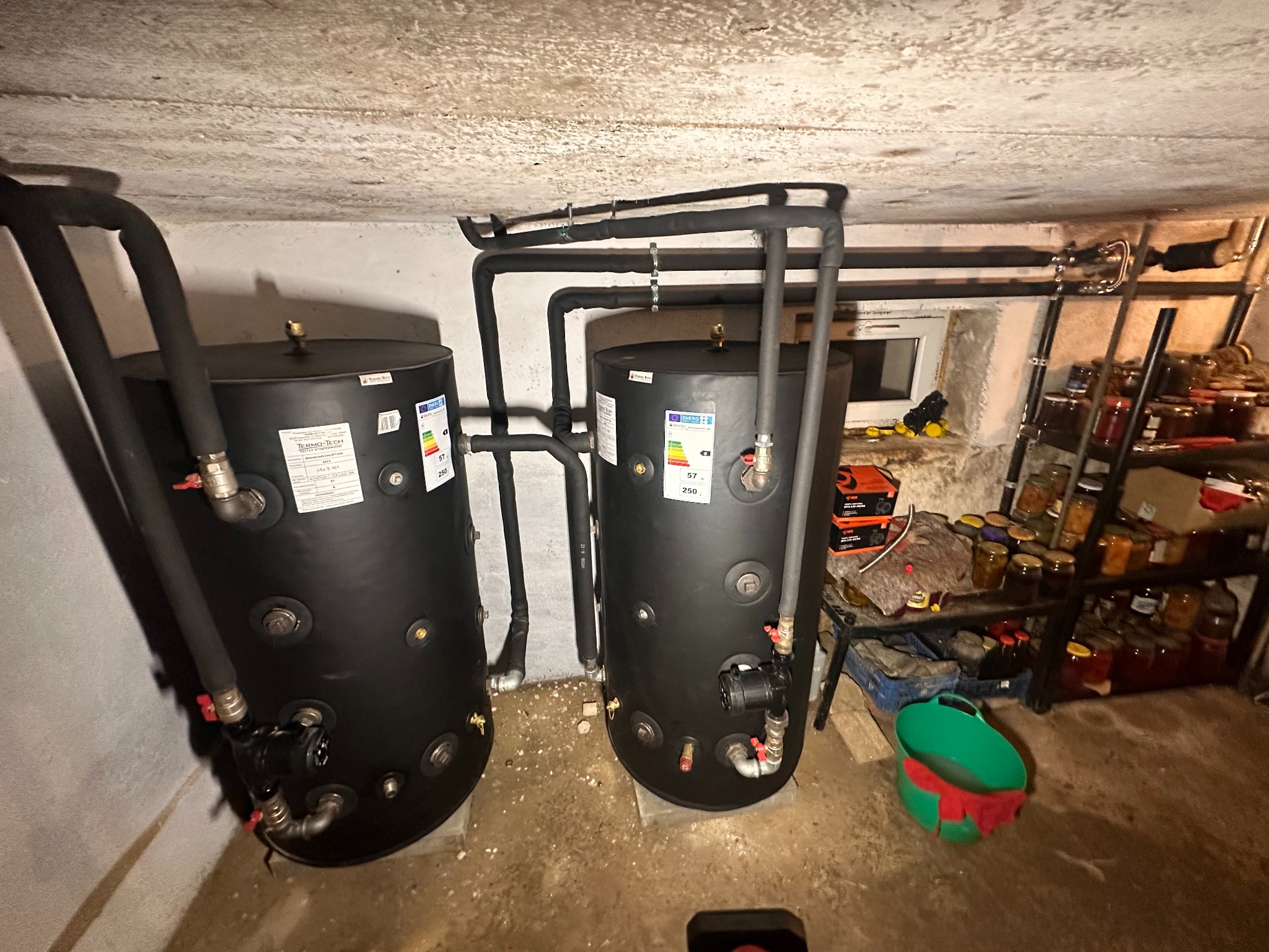 Dwa czarne zbiorniki buforowe z rurami w piwnicy, widoczne etykiety energetyczne i instalacja hydrauliczna. W tle okno i regał z przetworami.