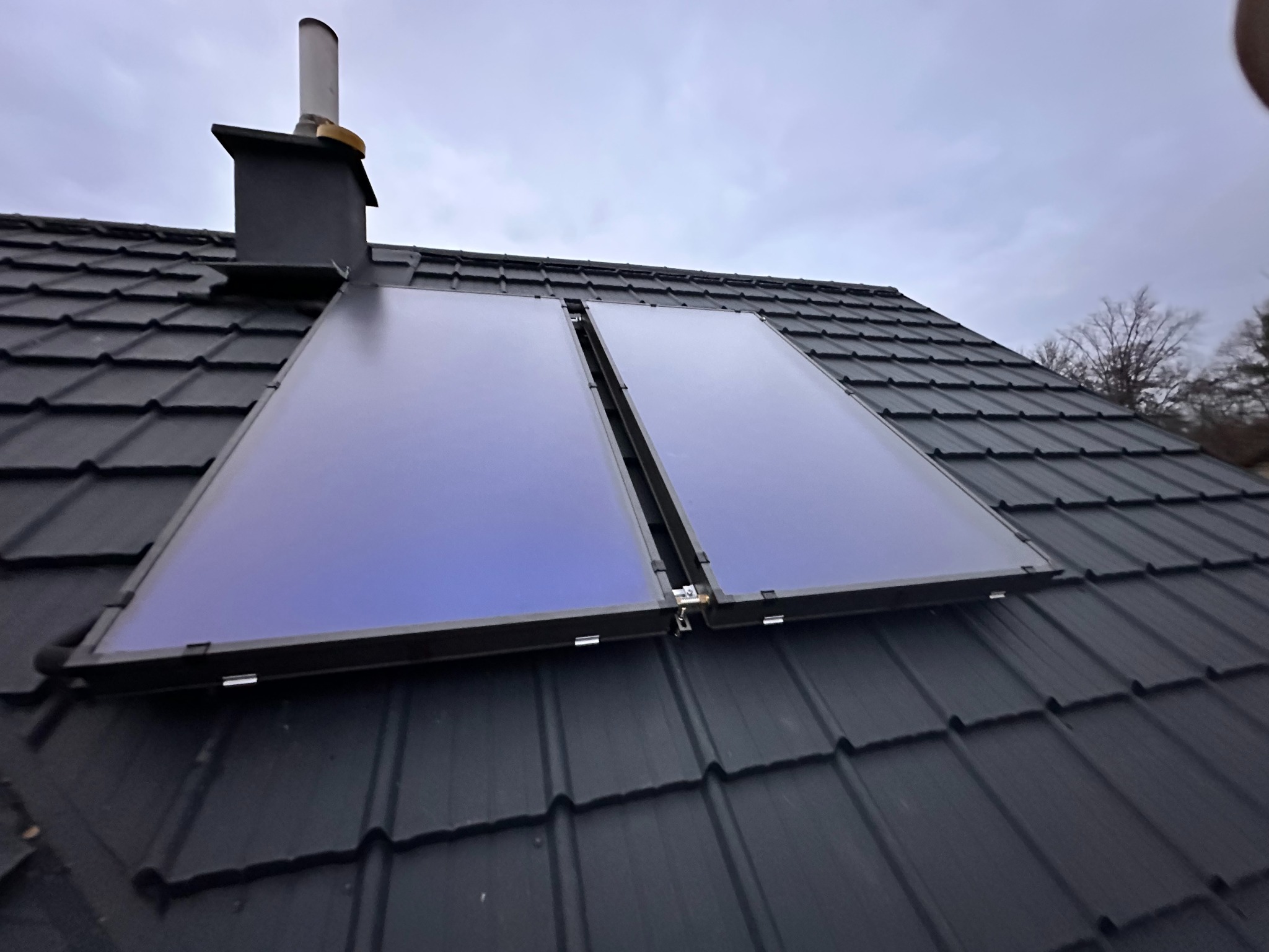 Dwa panele słoneczne zamontowane na ciemnym, spadzistym dachu z dachówki, widoczne z dołu pod kątem. W tle komin i fragmenty drzew.