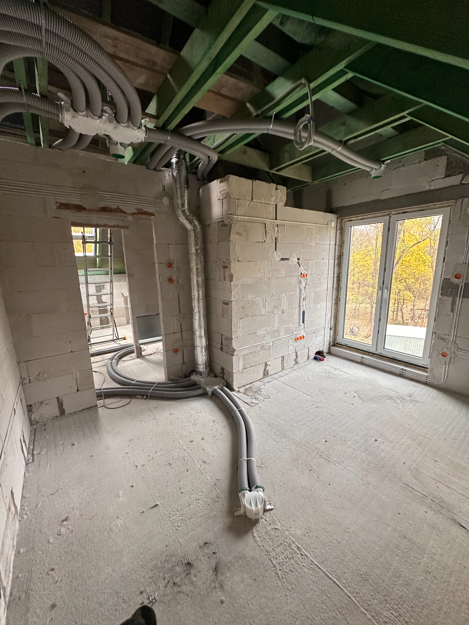 Surowy stan: Instalacja przewodów wentylacyjnych i elektrycznych w budynku z pustaków, widoczne okno z jesiennym krajobrazem. Szare rury biegną po ścianach i suficie.