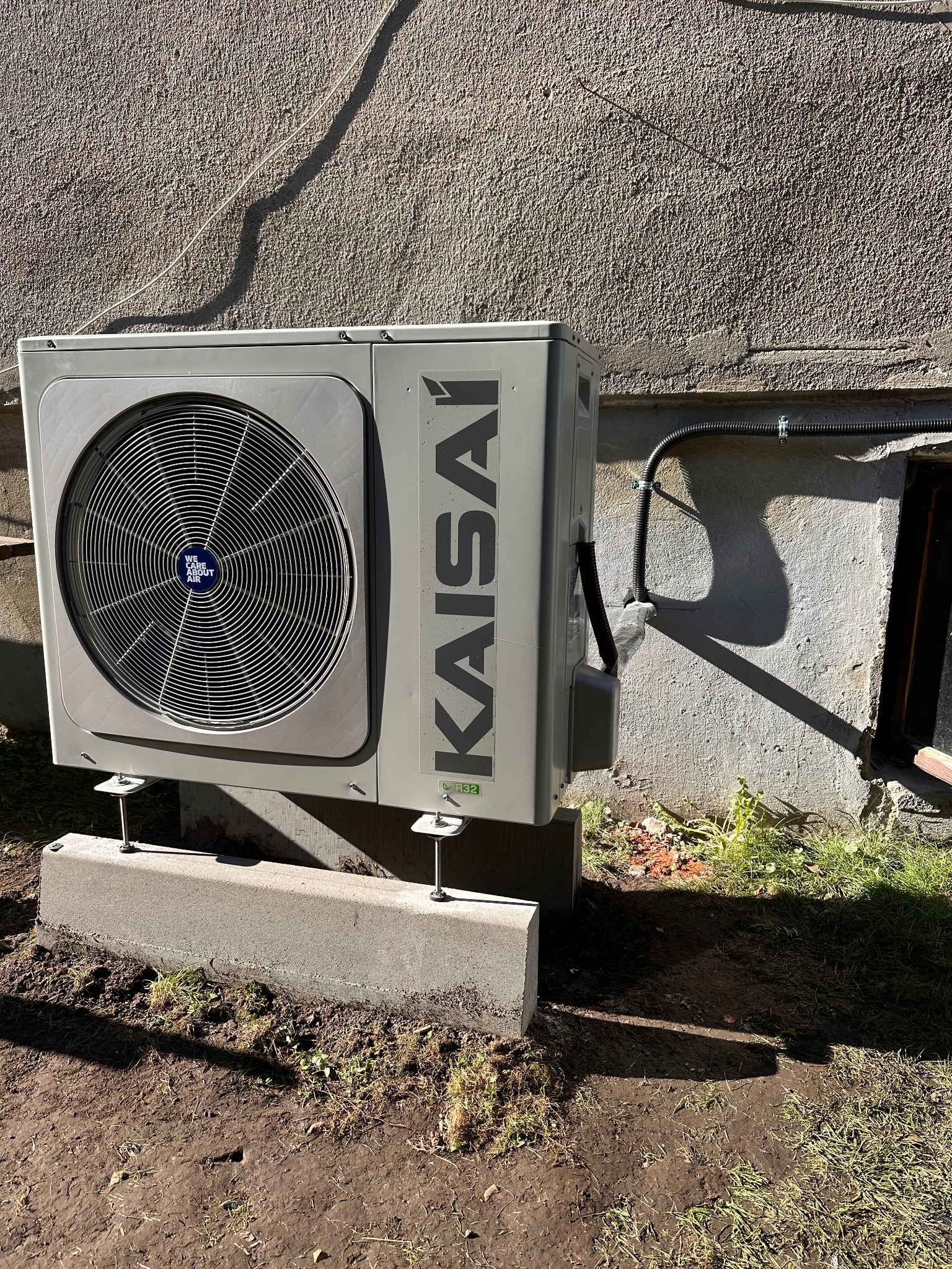 Szara jednostka zewnętrzna klimatyzacji Kaisai zamontowana na betonowych podstawach przy ścianie budynku, z widocznym przewodem elektrycznym.