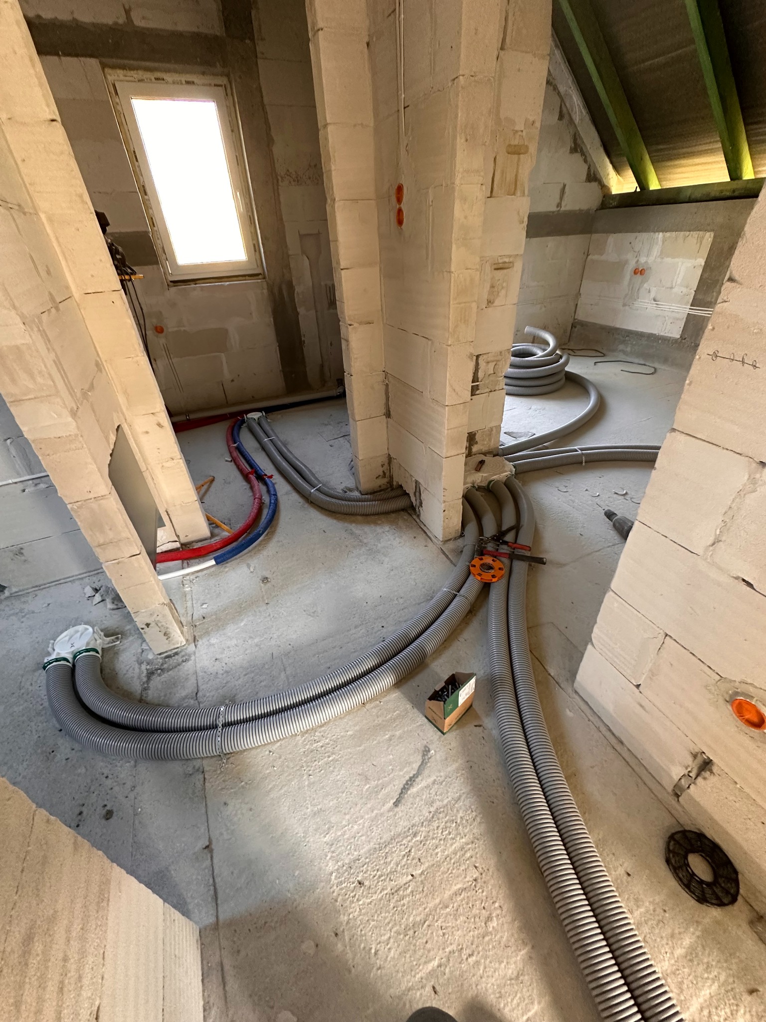 Wnętrze w trakcie instalacji: rury w peszlach prowadzone wzdłuż ścian z betonu komórkowego, widoczne narzędzia i okablowanie. Surowy stan, poddasze.