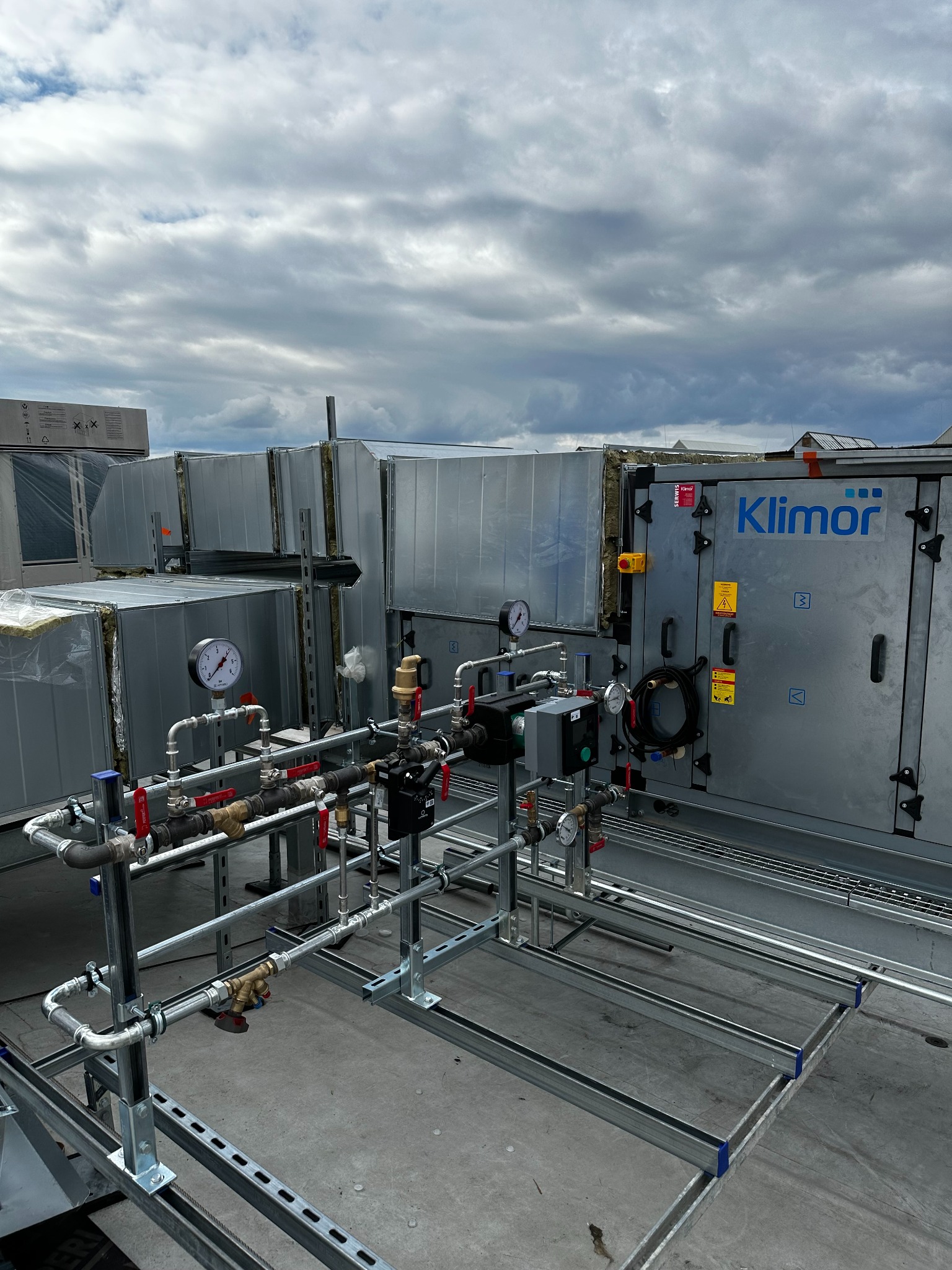 Instalacja przemysłowej klimatyzacji Klimor na dachu budynku, widoczne rury, zawory i manometry. Szare niebo w tle. Metalowe kanały wentylacyjne.