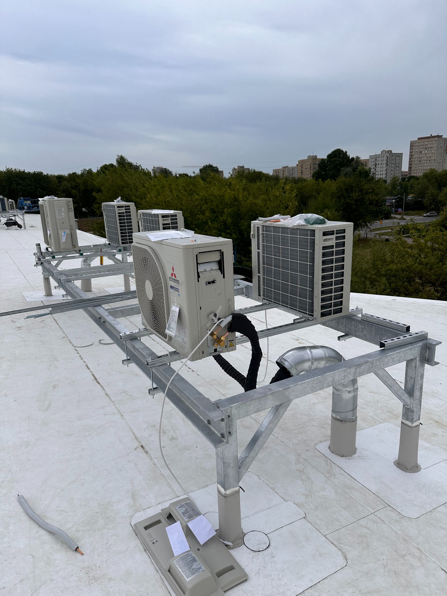 Instalacja klimatyzacji na dachu budynku. Widoczne jednostki zewnętrzne klimatyzatorów Mitsubishi na metalowej konstrukcji wsporczej, z przewodami i elementami montażowymi.