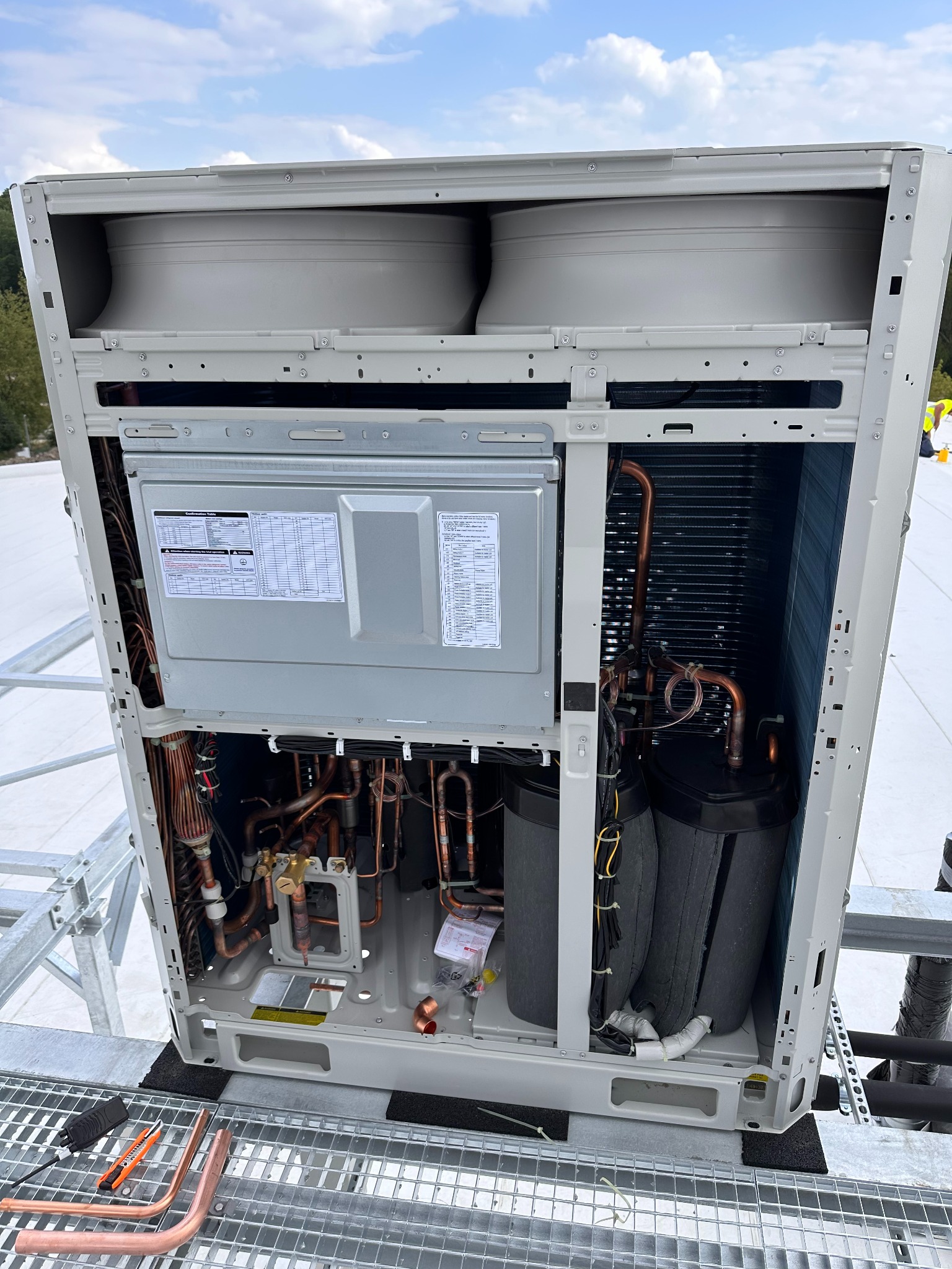 Wnętrze klimatyzatora z widocznymi miedzianymi rurami, wentylatorami i elementami izolacyjnymi. Na zewnątrz widoczne niebieskie niebo i fragment dachu.