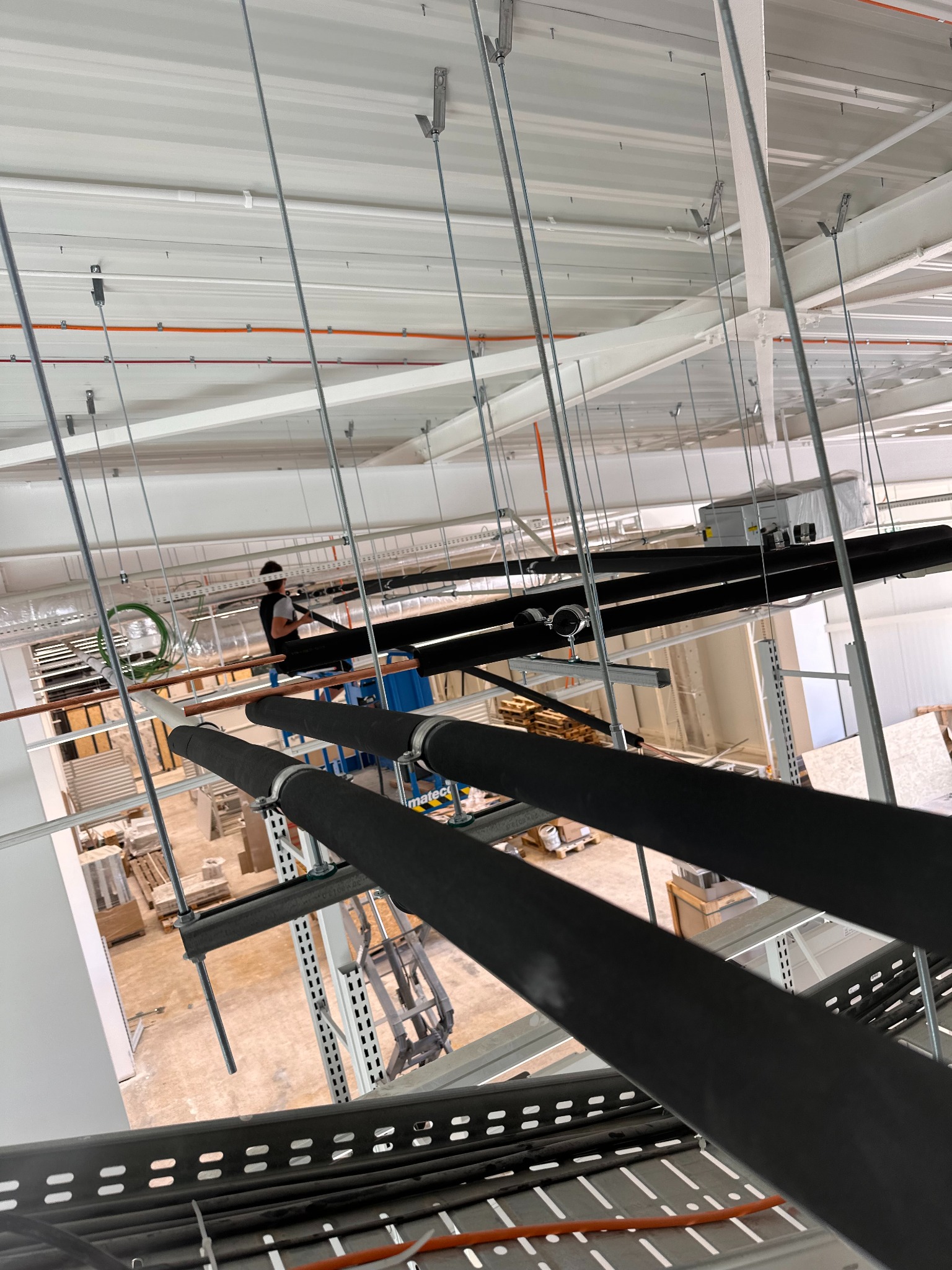 Montaż izolowanych rur w hali przemysłowej z widocznym pracownikiem na podnośniku, instalacja prowadzona pod sufitem, widok z dołu, oświetlenie dzienne.