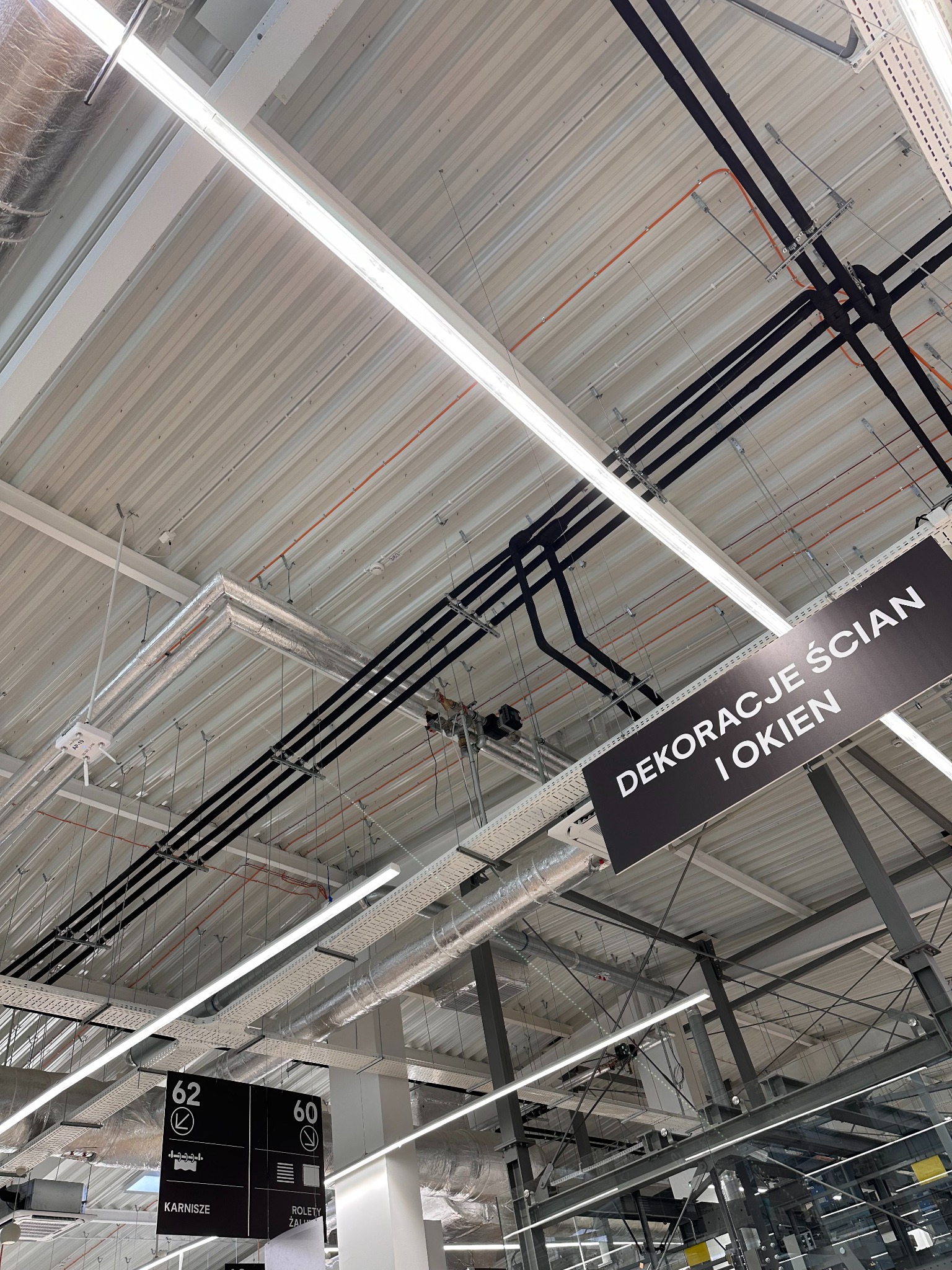Sufit hali z widocznymi instalacjami: rury wentylacyjne w srebrnej izolacji, czarne rury, oświetlenie liniowe i tabliczki informacyjne. Ujęcie z dołu pod kątem.