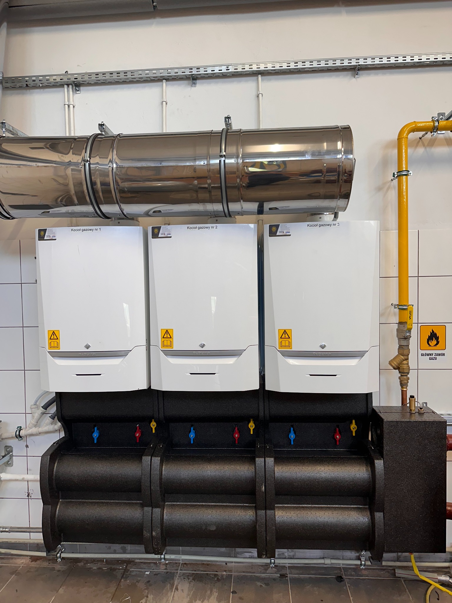 Trzy kotły gazowe De Dietrich, każdy z etykietą, zamontowane na czarnej podstawie z kolorowymi zaworami, z żółtą rurą gazową i oznaczeniem bezpieczeństwa.