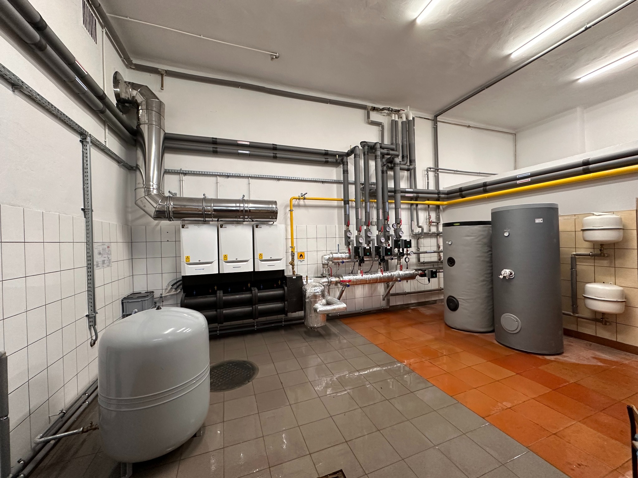 Zrealizowana instalacja grzewcza w kotłowni z dwoma szarymi zbiornikami, trzema białymi piecami i licznymi rurami. Podłoga wyłożona płytkami w dwóch kolorach.