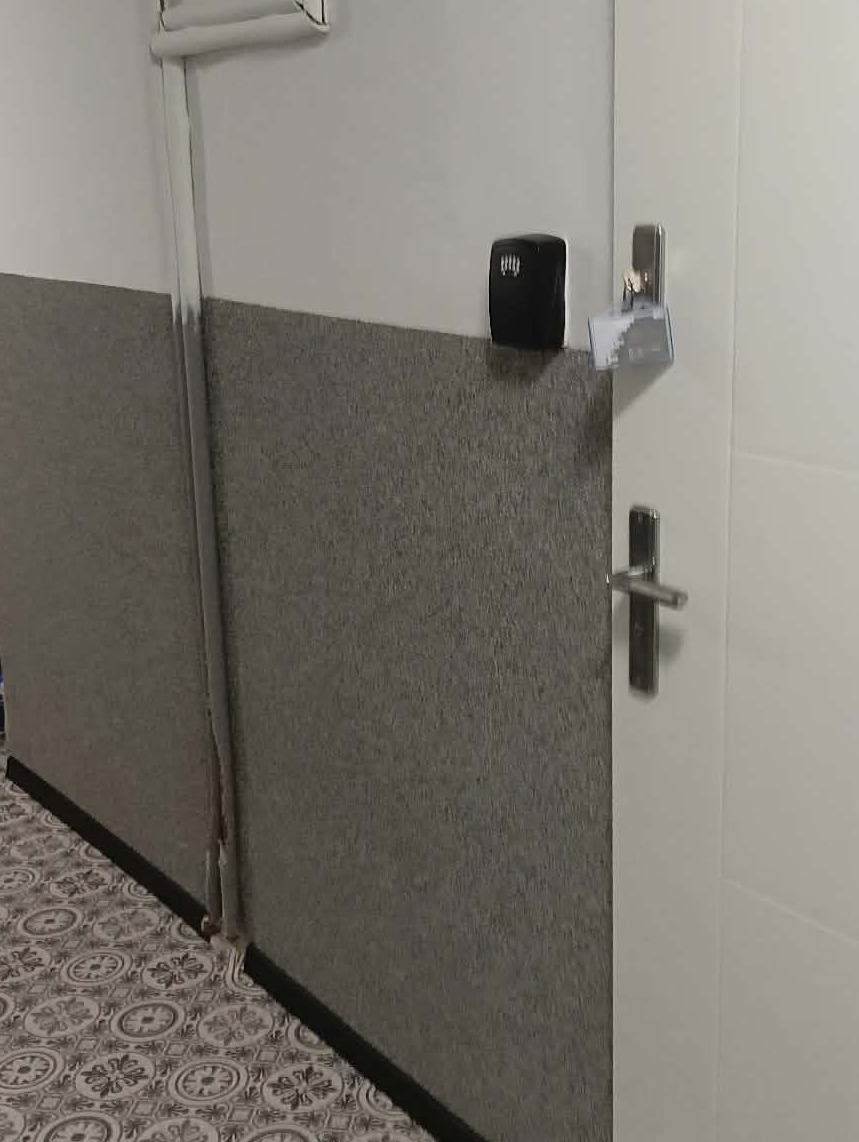 Szara, teksturowana ściana z listwą przypodłogową, białe drzwi z klamką i czarny głośnik na ścianie. W tle widoczna podłoga z wzorem.