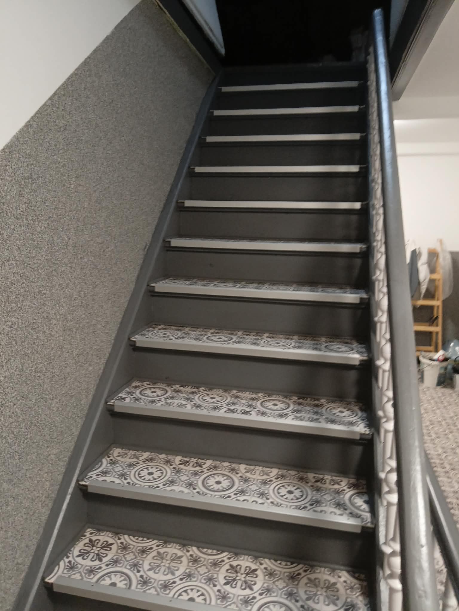 Schody po remoncie z szarymi stopniami i wzorzystymi płytkami. Widok z dołu, ukazujący odnowione wnętrze z elementami dekoracyjnymi i świeżym malowaniem.