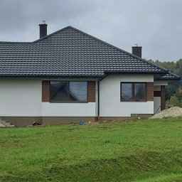 DAWBUD - Biały dom z ciemnym dachem na tle jesiennego lasu. Widoczne okna z brązowymi ramami i fragment trawnika. Dom po wykonaniu elewacji.