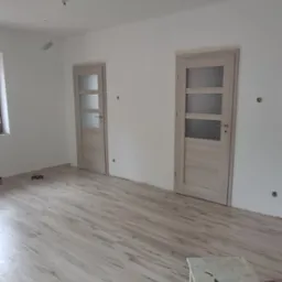 Jasne panele podłogowe w nowym pokoju z dwiema zamontowanymi, przeszklonymi drzwiami. Na pierwszym planie stołek z młotkiem. Świeżo po remoncie, gotowe do umeblowania.