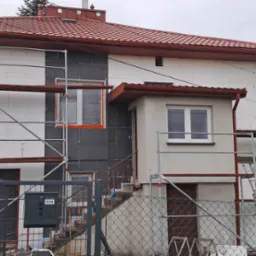 Dwukondygnacyjny dom w trakcie ocieplania. Część elewacji pokryta styropianem, część z szarym materiałem izolacyjnym. Rusztowania wokół budynku. Skrzynka pocztowa z numerem 559.