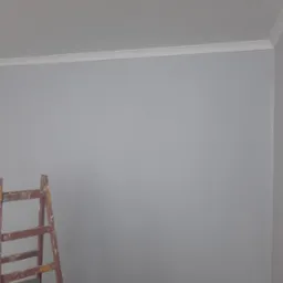 Świeżo pomalowana szara ściana z białym wykończeniem sufitu. W rogu drabina malarska, podłoga zabezpieczona folią. Widoczne pomarańczowe kliny dystansowe.