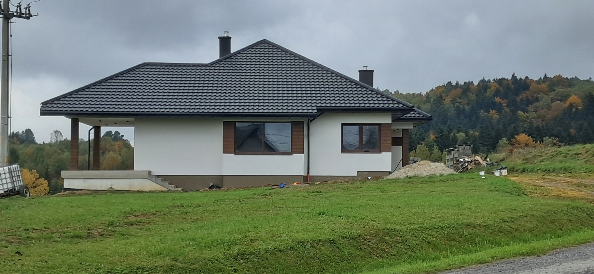 Biały dom z ciemnym dachem na tle jesiennego lasu. Widoczne okna z brązowymi ramami i fragment trawnika. Dom po wykonaniu elewacji.