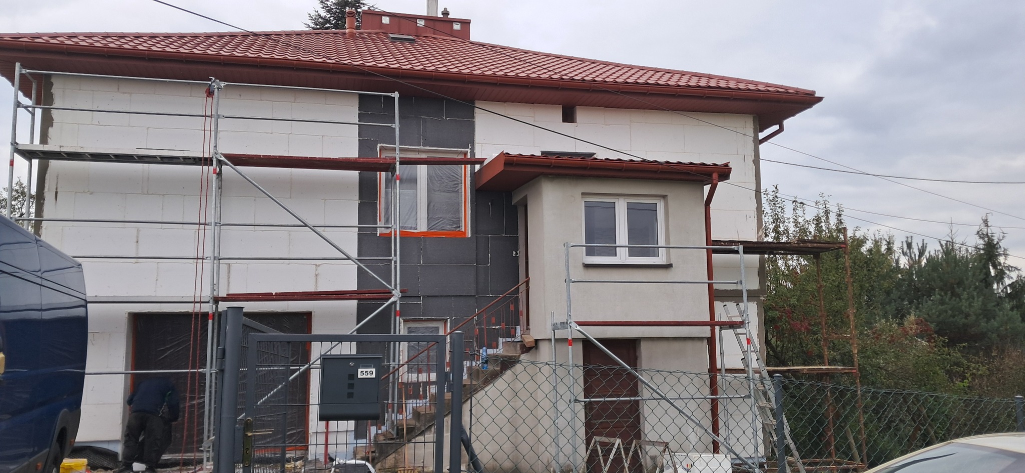 Dwukondygnacyjny dom w trakcie ocieplania. Część elewacji pokryta styropianem, część z szarym materiałem izolacyjnym. Rusztowania wokół budynku. Skrzynka pocztowa z numerem 559.