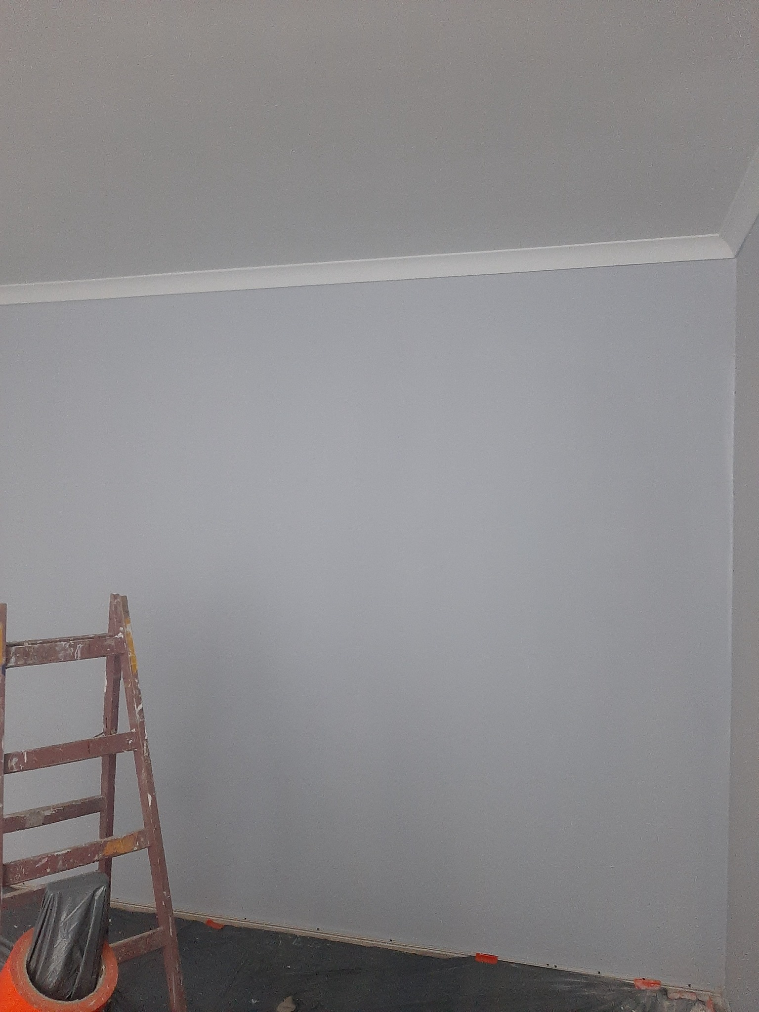Świeżo pomalowana szara ściana z białym wykończeniem sufitu. W rogu drabina malarska, podłoga zabezpieczona folią. Widoczne pomarańczowe kliny dystansowe.