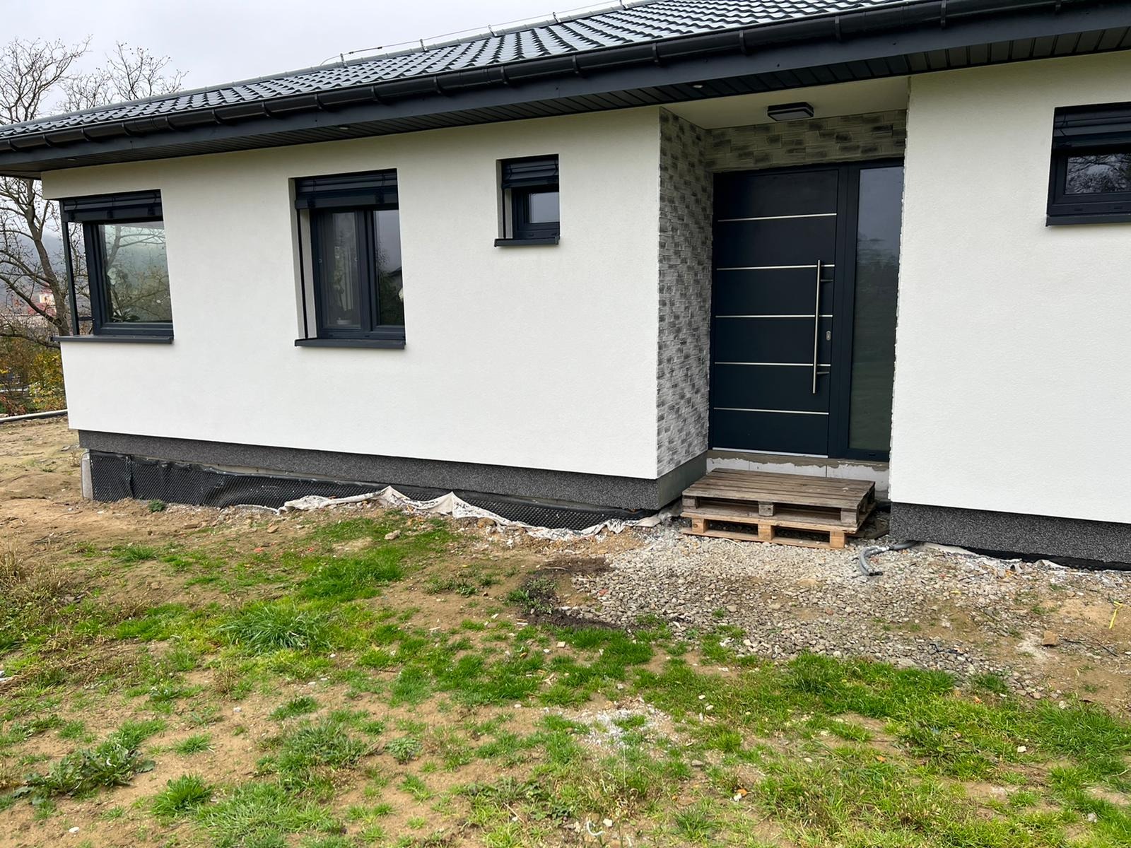 Nowoczesny dom jednorodzinny z białą elewacją, grafitową stolarką okienną i drzwiami wejściowymi. Wykończenie elewacji imitujące kamień przy wejściu. Teren wokół domu w trakcie zagospodarowania.