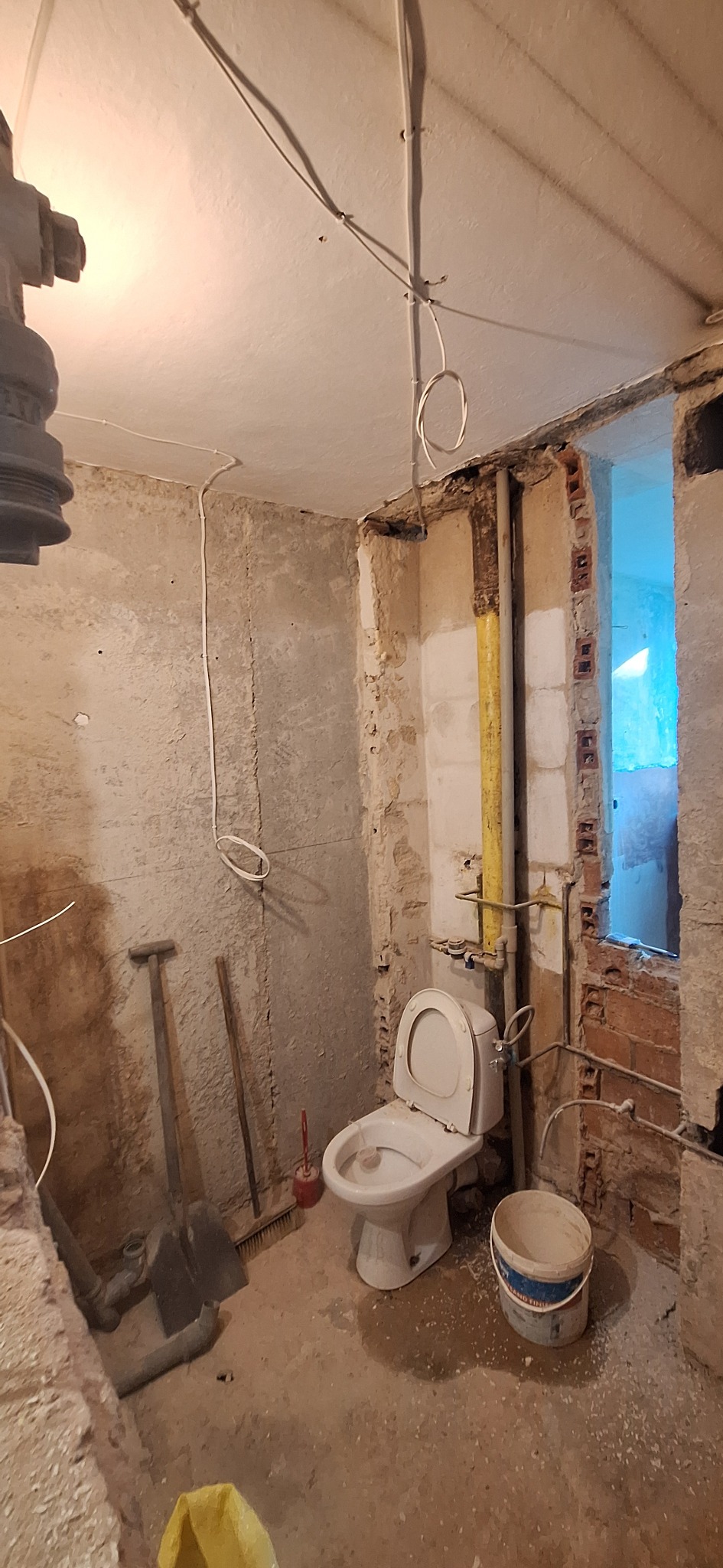 Łazienka w trakcie remontu: surowe ściany, instalacje, toaleta, narzędzia. Widoczne przewody elektryczne i rury. Okno z widokiem na zewnątrz.