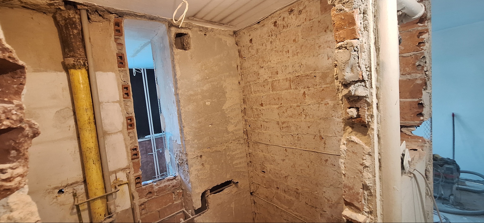 Wnętrze łazienki w trakcie remontu: odsłonięte cegły, fragmenty instalacji, surowe ściany z resztkami tynku. Widoczne rury i okno z kratą.