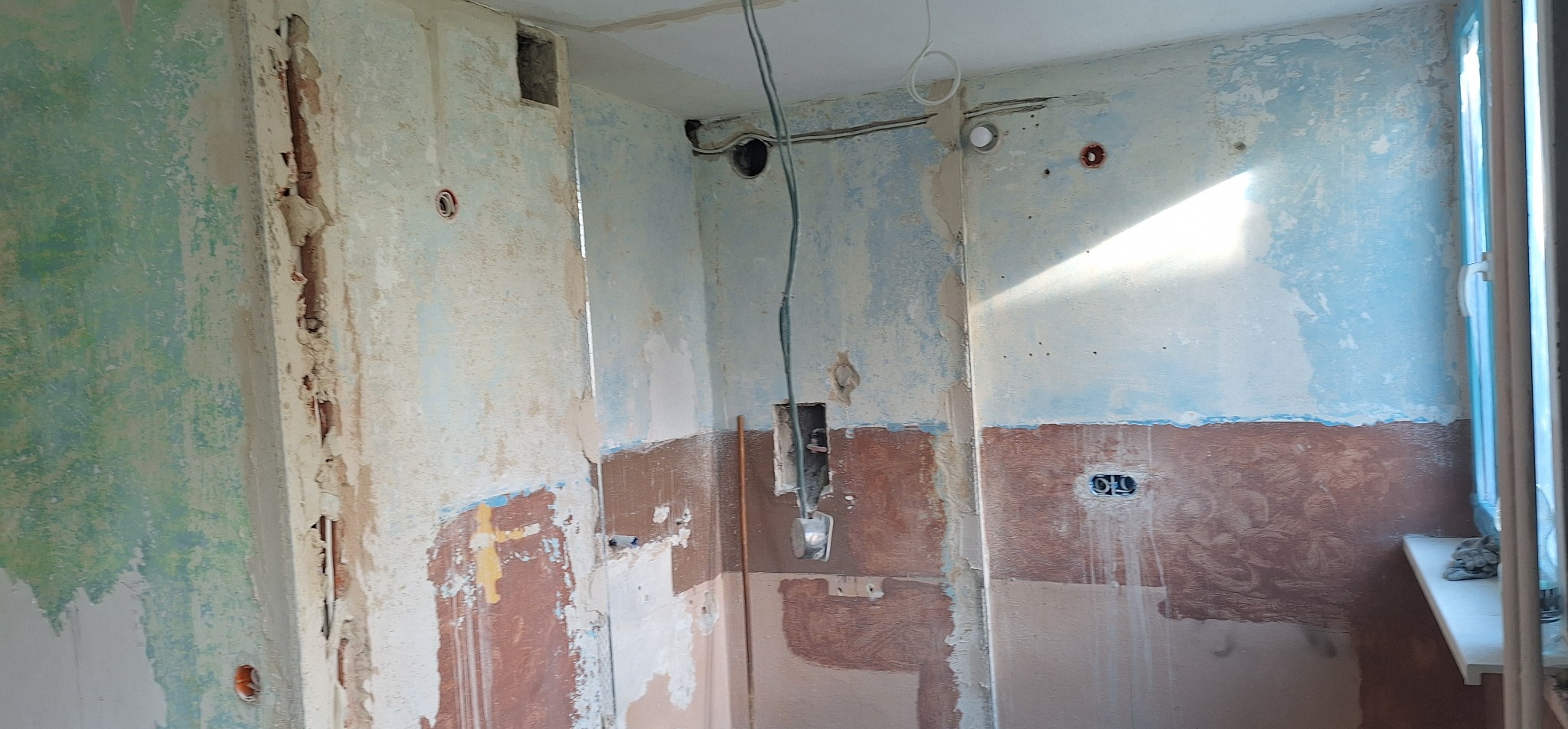 Wnętrze w trakcie remontu: ściany z resztkami farby, odsłonięte instalacje elektryczne i fragment okna. Widoczne ślady po starych tapetach i przygotowanie do dalszych prac wykończeniowych.