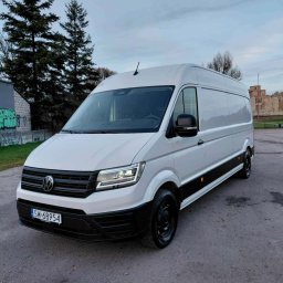 Michał Wojdas Usługi Transportowo - Kurierskie - Biały, wysoki bus Volkswagen Crafter na parkingu, z widoczną tablicą rejestracyjną SW-68954, na tle drzew i budynku przypominającego zamek.