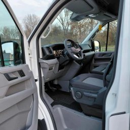 Michał Wojdas Usługi Transportowo - Kurierskie - Wnętrze białego busa z otwartymi drzwiami kierowcy, widok na deskę rozdzielczą, kierownicę i szare fotele. Widoczne drzewa w tle.
