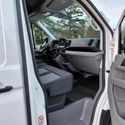 Michał Wojdas Usługi Transportowo - Kurierskie - Wnętrze białego busa z otwartymi drzwiami, widoczne szare fotele, deska rozdzielcza z ekranem dotykowym i gumowa wycieraczka. Widok z zewnątrz na kabinę pojazdu.