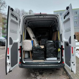 Michał Wojdas Usługi Transportowo - Kurierskie - Wnętrze białego busa z otwartymi drzwiami, wypełnione meblami i kartonami zabezpieczonymi czarną folią, gotowymi do transportu. Widok z tyłu pojazdu.