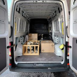 Michał Wojdas Usługi Transportowo - Kurierskie - Wnętrze białego busa z otwartymi drzwiami, częściowo załadowane kartonami i drewnianą konstrukcją. Widok z tyłu pojazdu, na zewnątrz budynek z cegły.