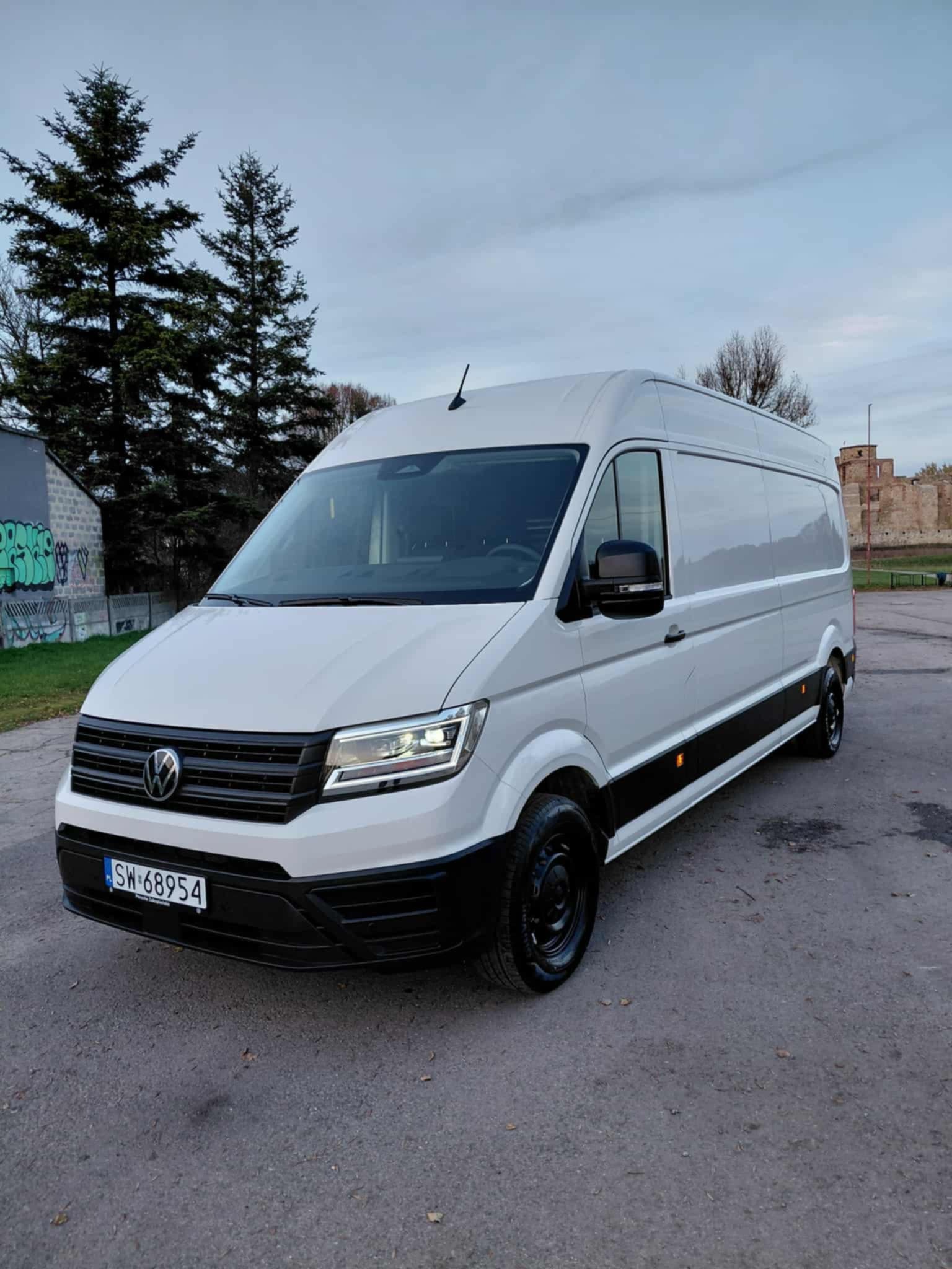 Biały bus Volkswagen Crafter z czarnymi elementami, zaparkowany na tle drzew i murów zamkowych. Tablica rejestracyjna widoczna.