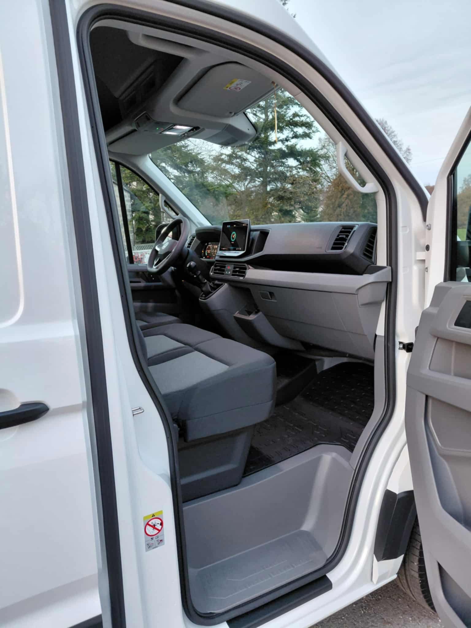 Wnętrze białego busa z otwartymi drzwiami, widok na deskę rozdzielczą, kierownicę i szare fotele. Na podłodze gumowe dywaniki. Widoczne drzewa w tle.