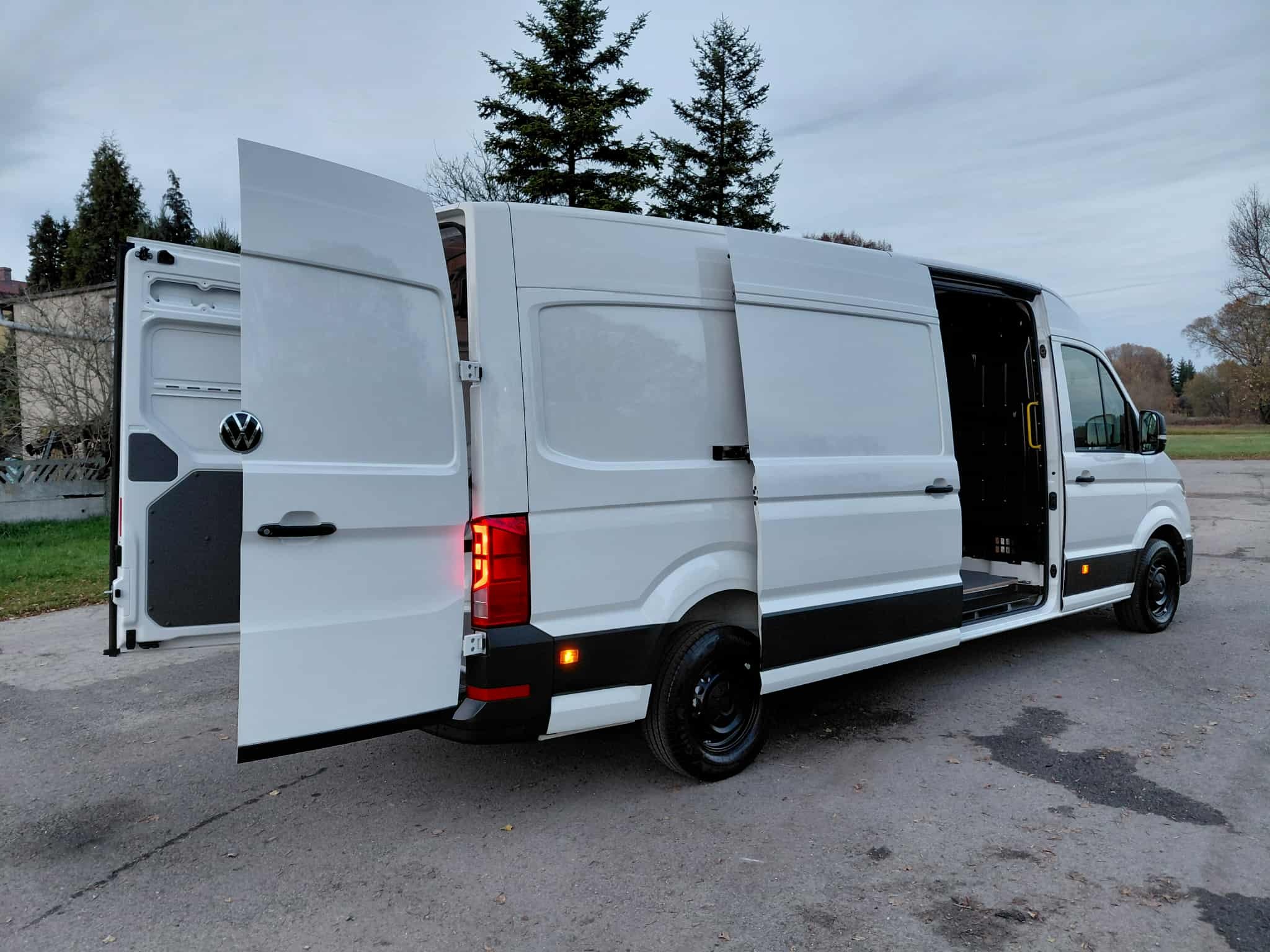 Biały bus dostawczy z otwartymi drzwiami bocznymi i tylnymi, zaparkowany na zewnątrz. Widoczne drzewa w tle. Ujęcie z perspektywy lekko od dołu.