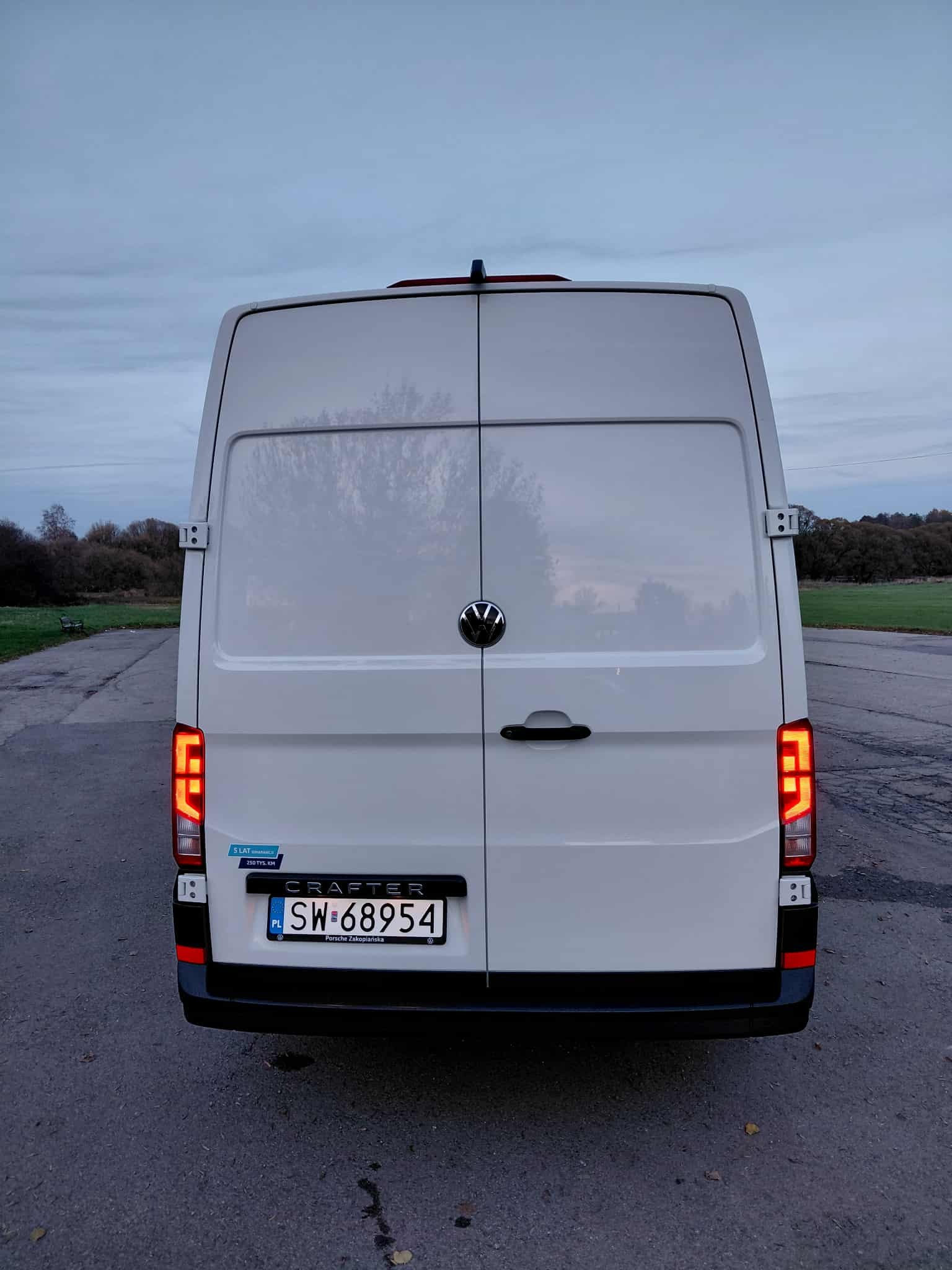 Tył białego busa VW Crafter z polską tablicą rejestracyjną SW-68954 na tle asfaltowej drogi i zieleni. Widoczne światła stopu i naklejka '5 lat gwarancji'.