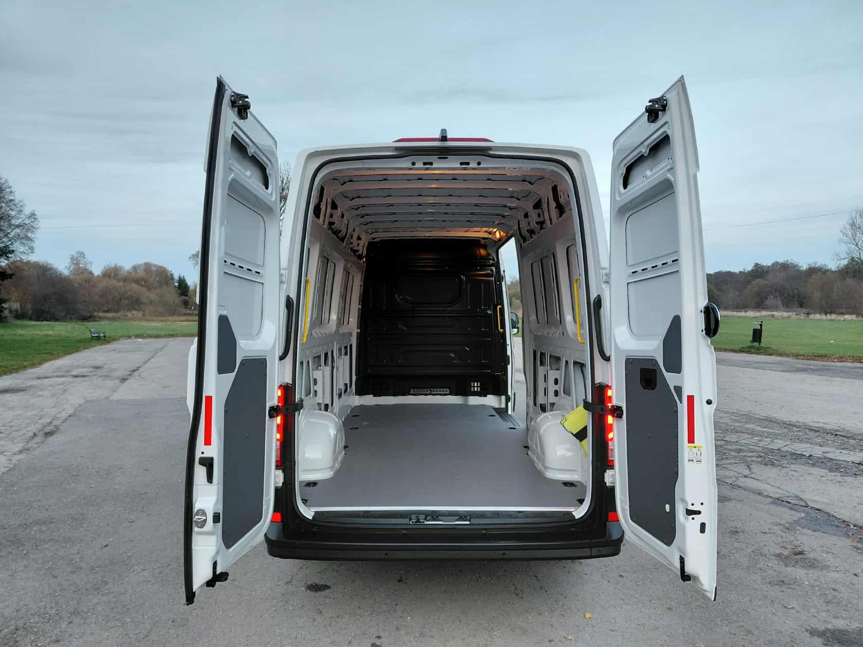 Wnętrze białego, pustego busa z otwartymi drzwiami, gotowego do załadunku. Widoczna przestrzeń ładunkowa i elementy mocujące. Tło stanowi krajobraz z drzewami i szarym niebem.