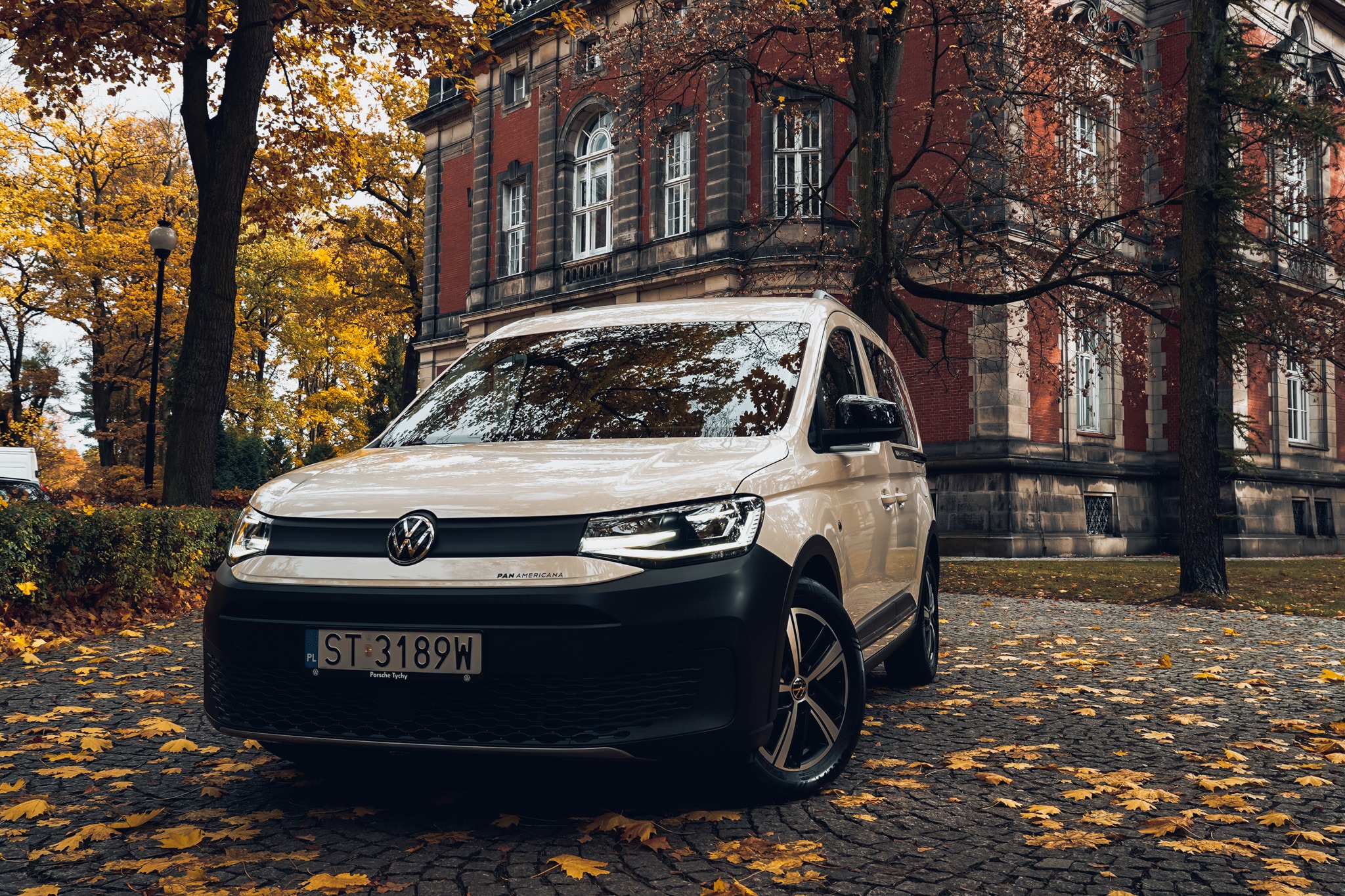 Kremowy Volkswagen Caddy PanAmericana na tle jesiennego parku i ceglanego budynku. Liście na brukowanej drodze. Tablica rejestracyjna ST 3189W.