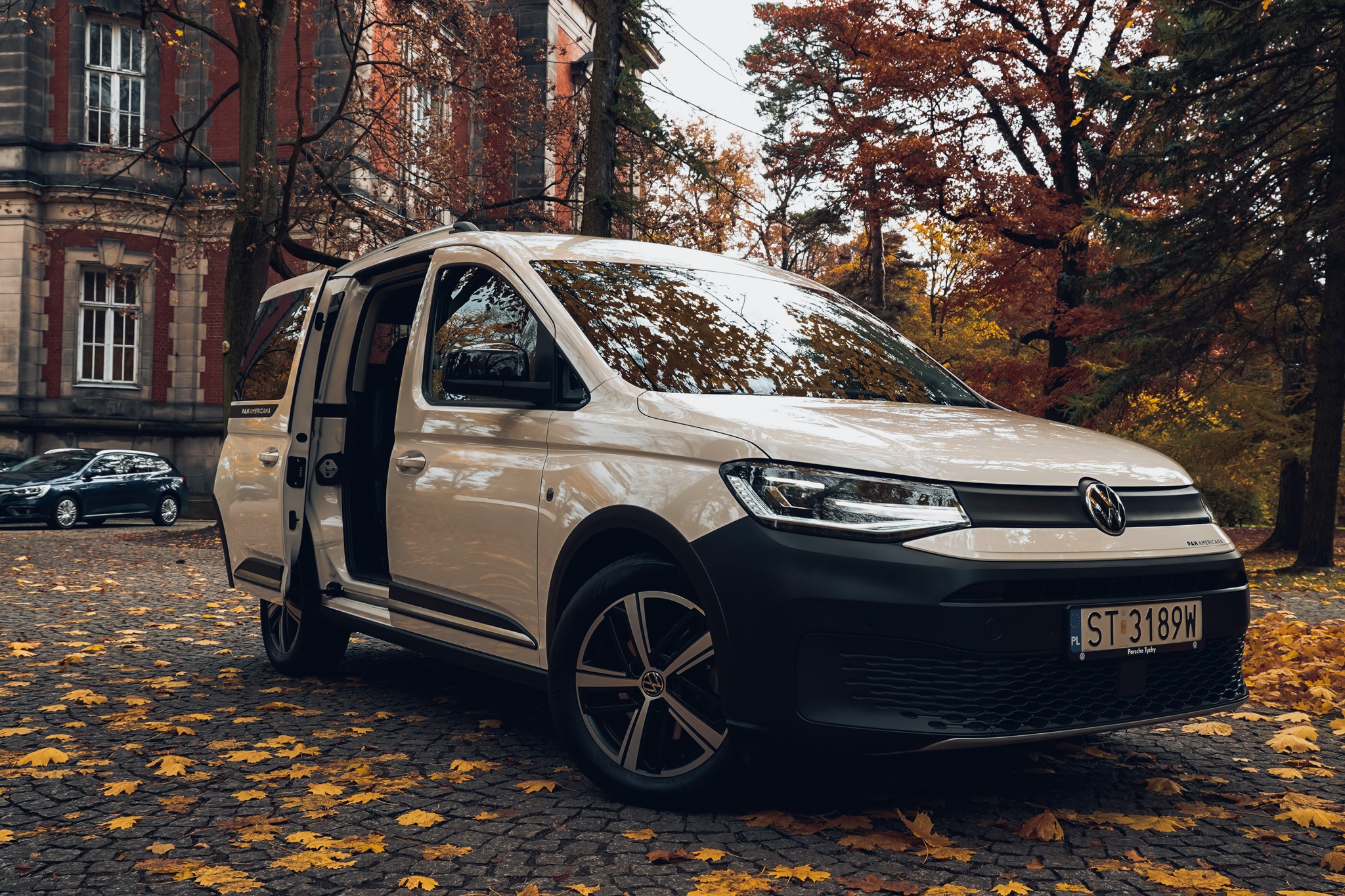 Kremowy Volkswagen Caddy PanAmericana z otwartymi drzwiami na tle jesiennego krajobrazu i ceglanego budynku. Na brukowanej drodze leżą opadłe liście.