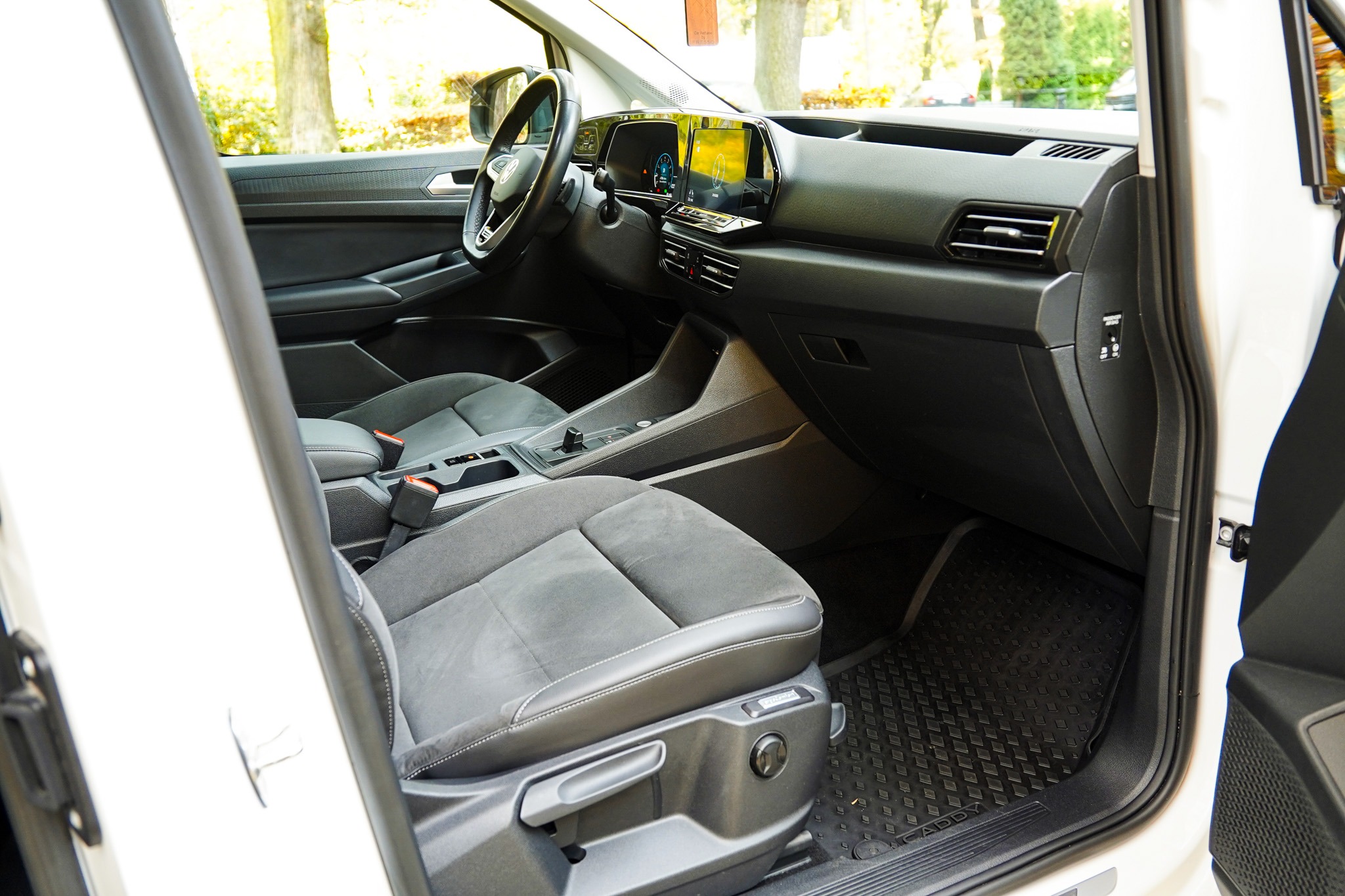 Wnętrze białego samochodu z szarą tapicerką, widok z otwartych drzwi kierowcy, deska rozdzielcza z ekranem dotykowym, czarne dywaniki z wzorem rombów.