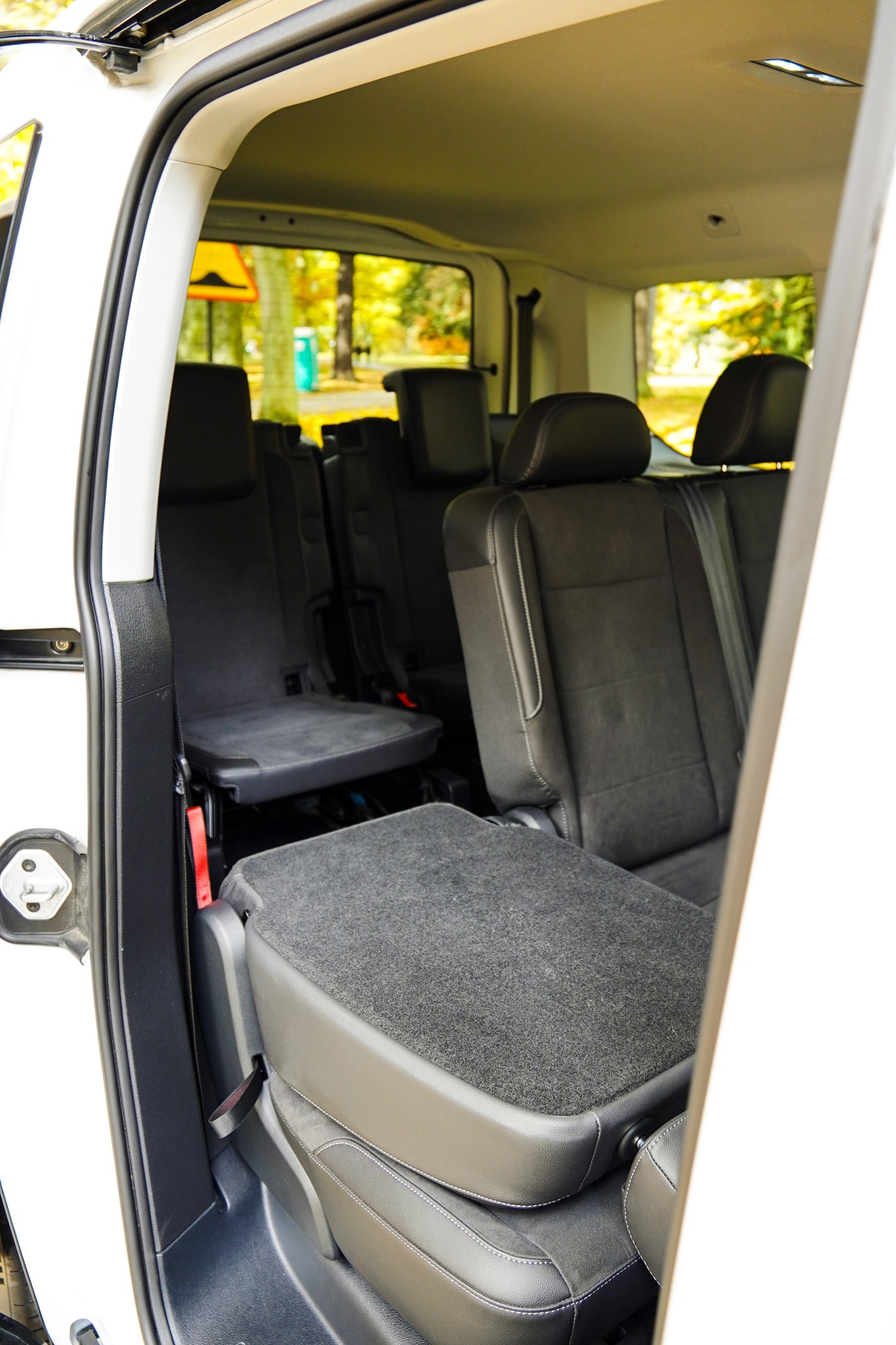 Wnętrze białego vana z widocznymi szarymi fotelami pasażerskimi i otwartymi drzwiami, przez które widać zieleń drzew.